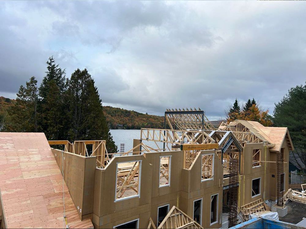 Une maison en construction, à ossature bois, face à un lac sous un ciel nuageux.