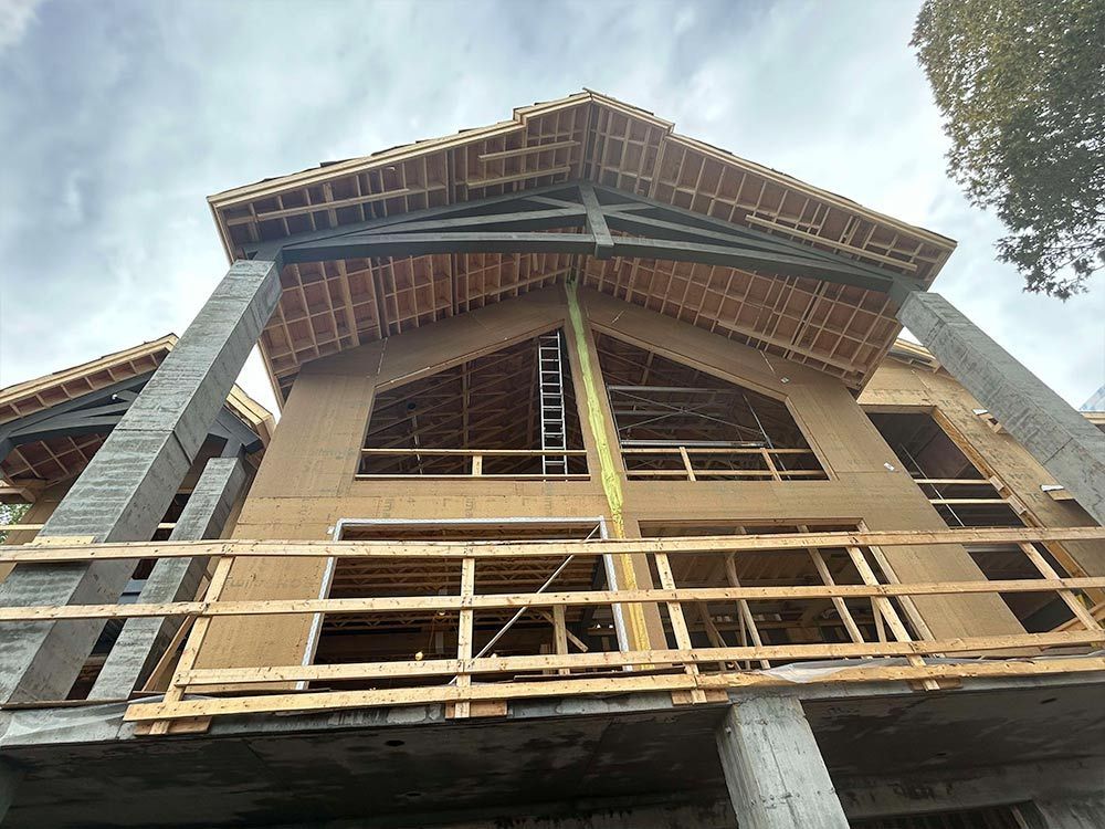 Construction d'un grand bâtiment en bois avec poutres et colonnes apparentes, et un toit inachevé, sous un ciel nuageux.