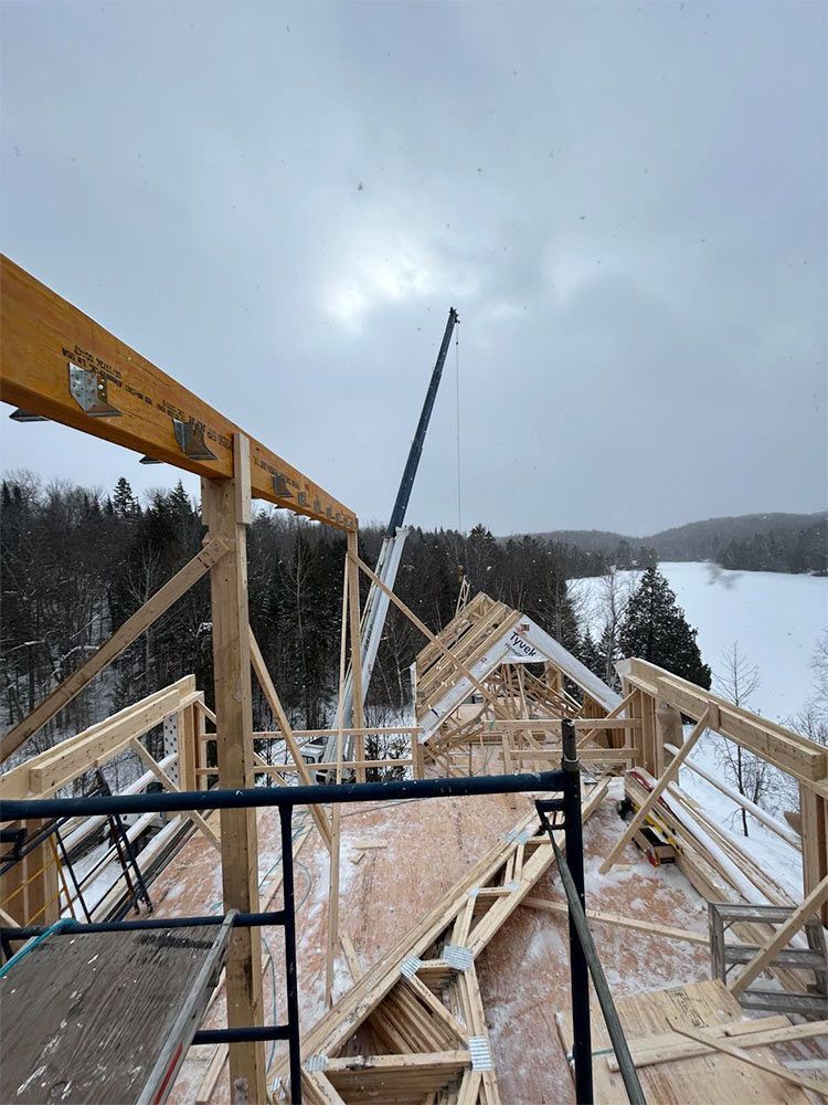 Chantier de construction : une grue soulève une ferme de toit et la pose sur une structure en bois dans un décor enneigé.