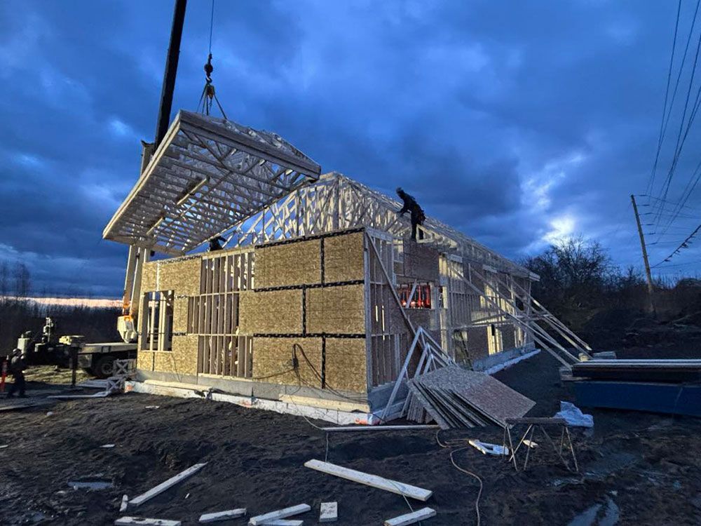 Construction d'un bâtiment. Une grue soulève une section de toiture au-dessus de la charpente en bois ; des ouvriers la fixent. Ciel du soir.