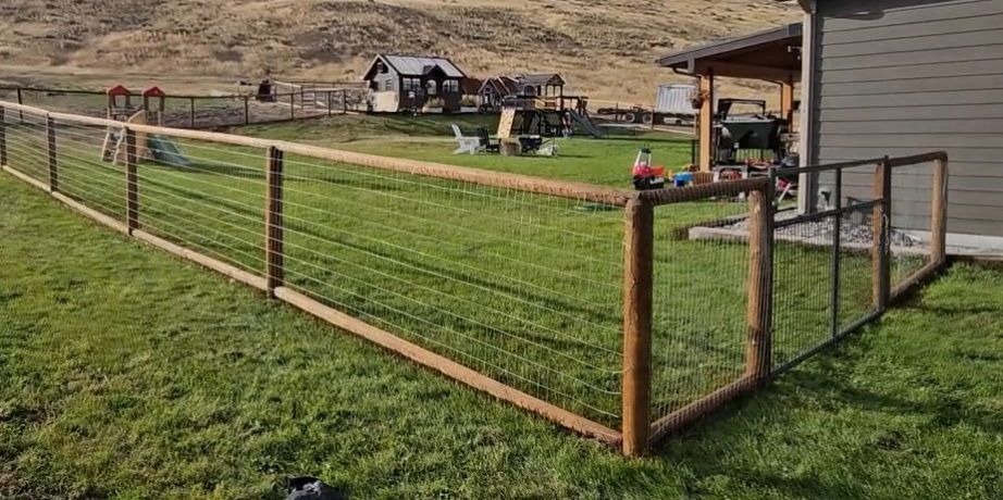 Wooden fence enclosing a grassy yard beside a gray house, with a shed and trailer in the background