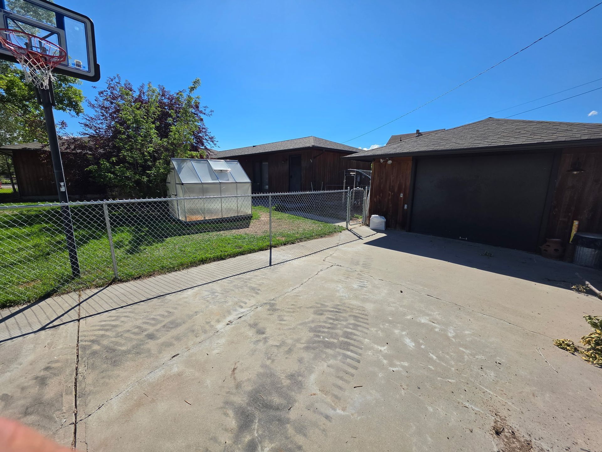 Sunny backyard driveway beside a wooden garage, chain-link fence, shed, and basketball hoop.