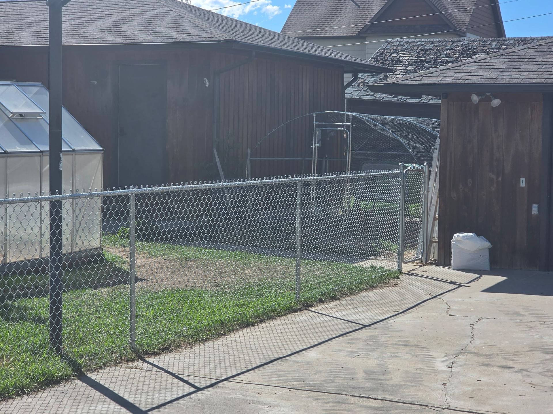 Chain-link fence along a driveway beside a wooden shed and houses on a sunny day