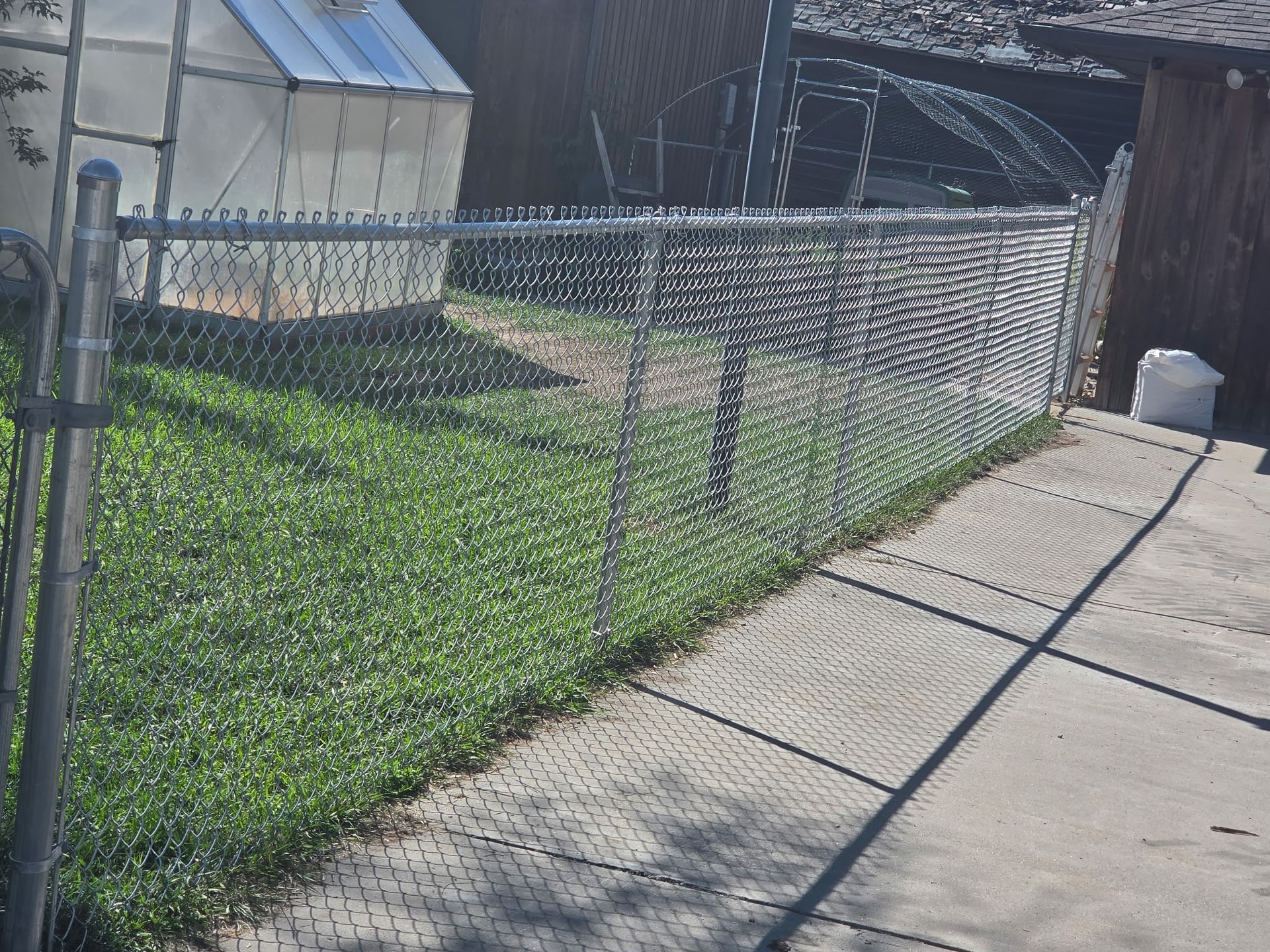 Chain-link fence along a concrete sidewalk beside a grassy yard and small shed