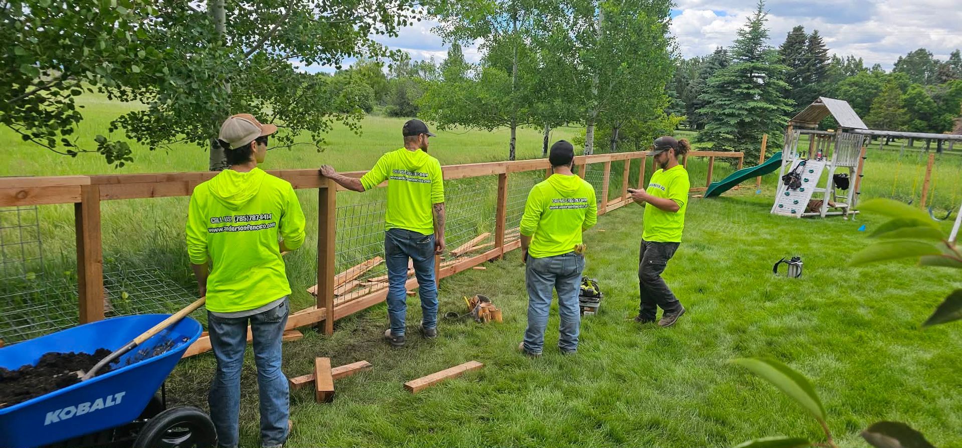 People in neon shirts working with shovels in a grassy garden near a blue wheelbarrow and small structure