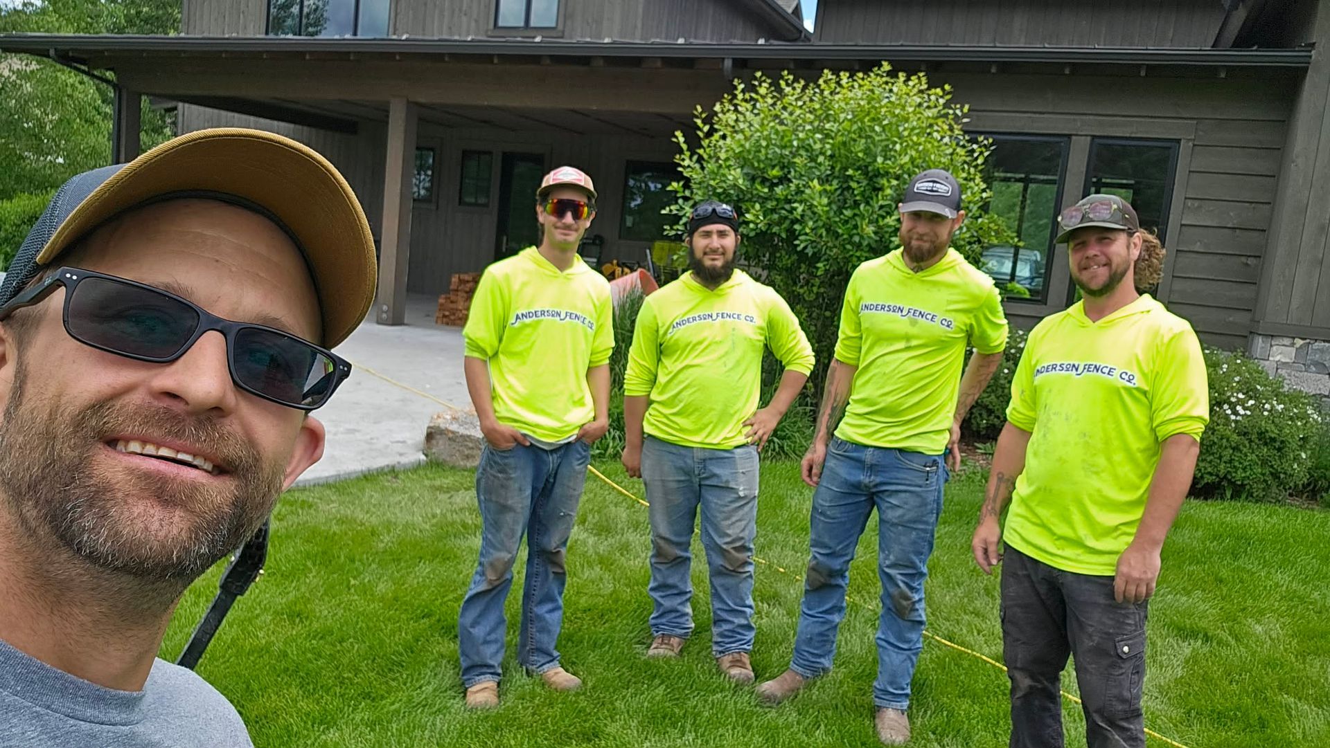 Five men in neon yellow shirts pose on a lawn outside a house, with one smiling selfie in the foreground.
