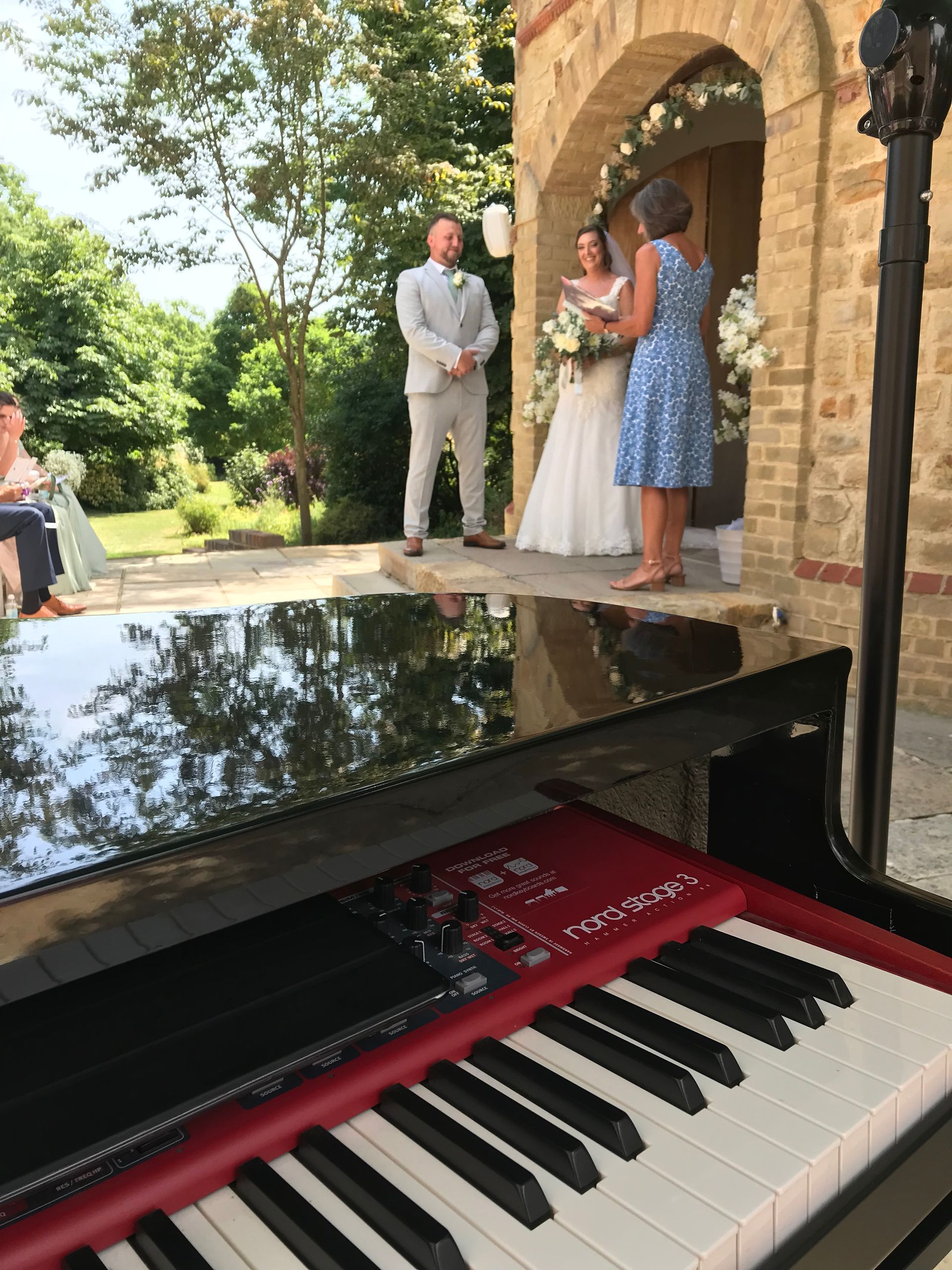 A piano in the foreground on a sunny day while a lady in a blue dress marries a bride and groom