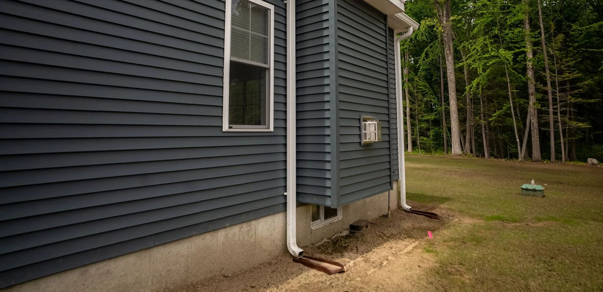 Blue house siding with a white downspout beside a grassy yard and trees