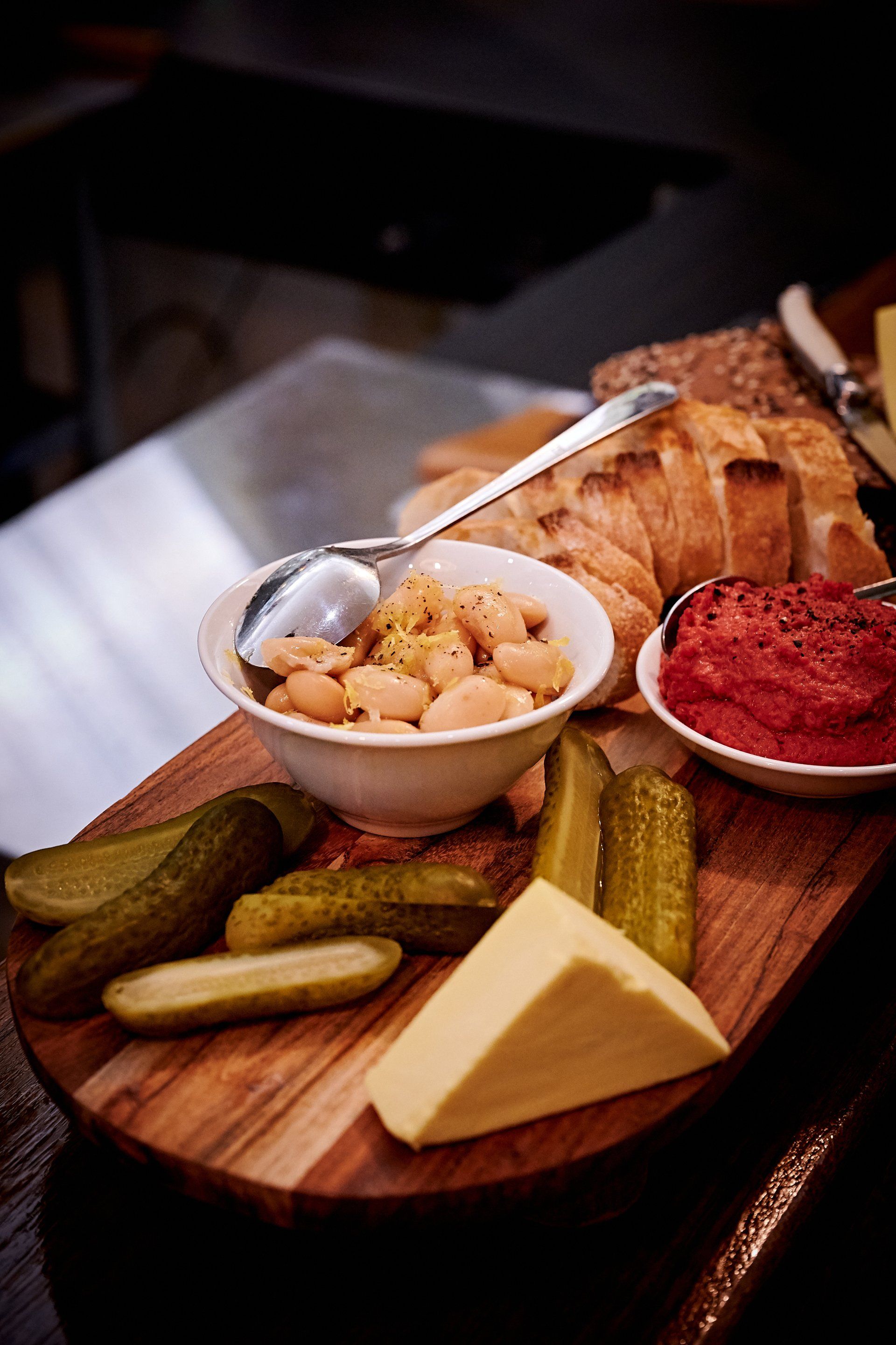Wooden board with white beans, pickles, cheese, bread, and red dip. Vegetarian.
