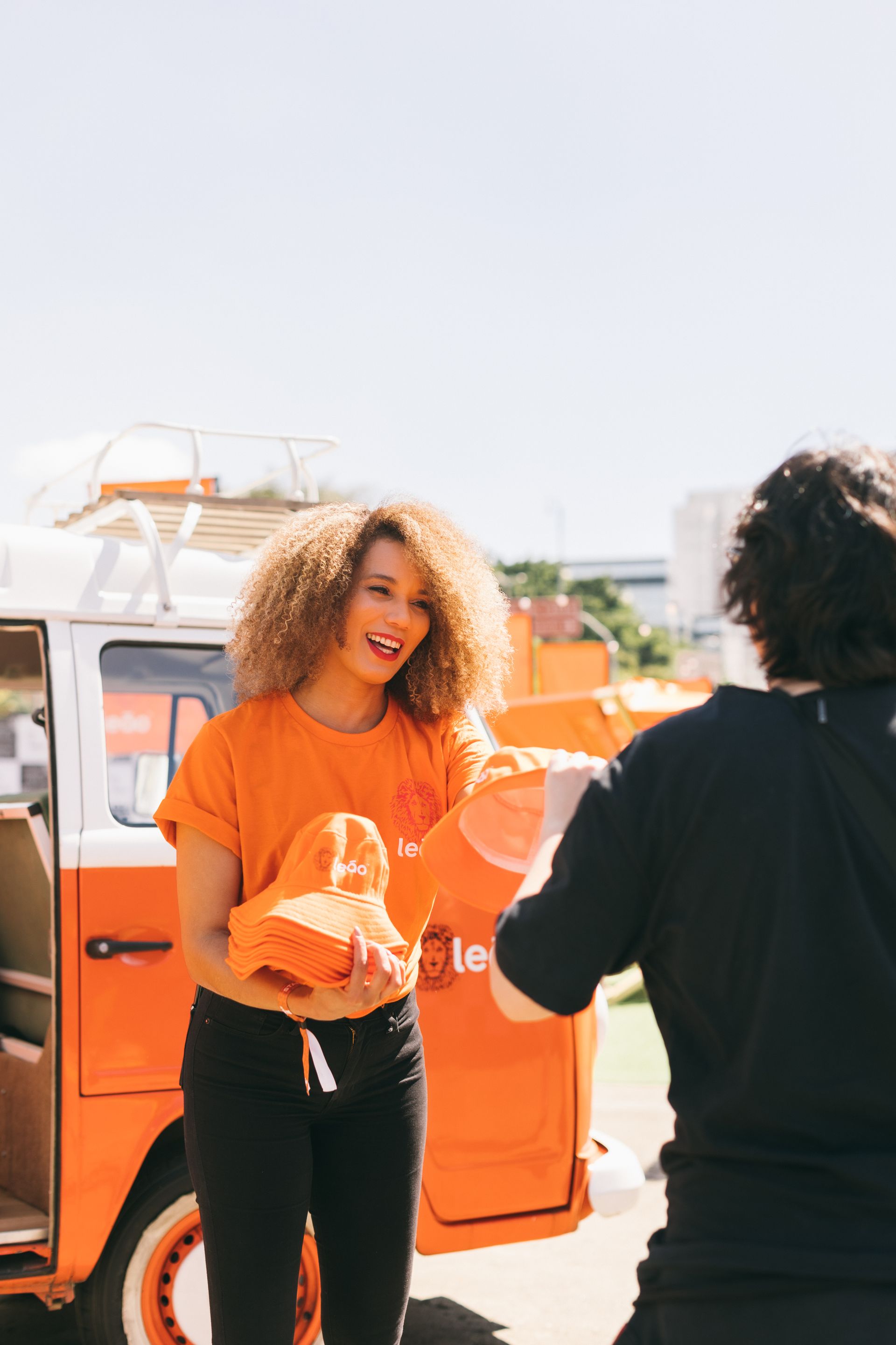 Uma mulher com uma camisa laranja está dando uma sacola para outra mulher em frente a uma van laranja.