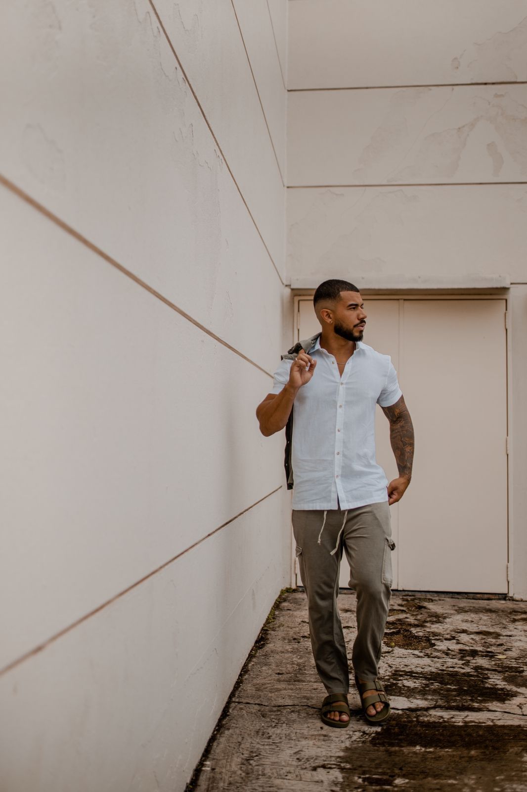 Um homem de camisa branca e calça cáqui está parado em frente a uma parede branca.