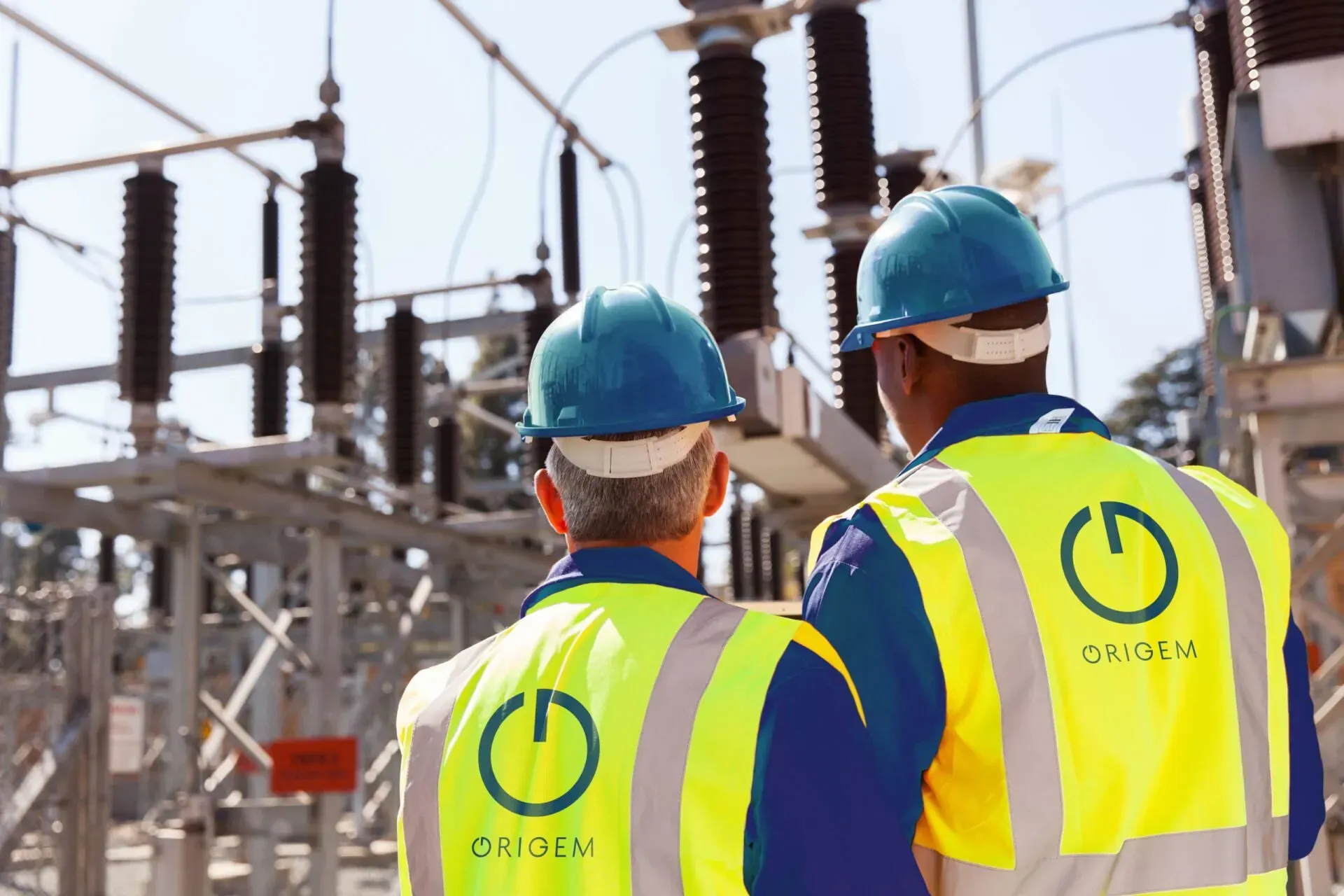 Dois homens usando capacetes e coletes de segurança estão parados em frente a uma usina de energia.