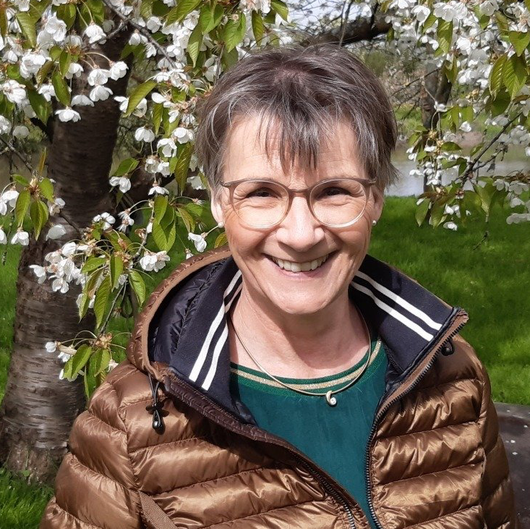 Eine lächelnde Person mit Brille und brauner Jacke steht im Freien an blühenden Baumzweigen.