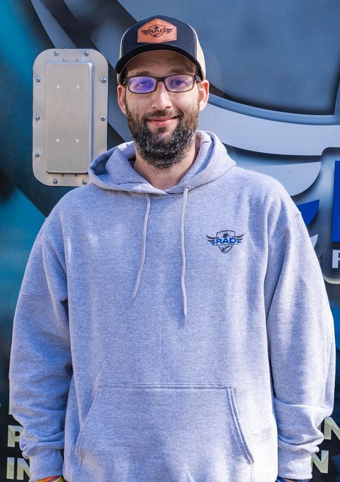 A man with a beard and glasses is wearing a grey hoodie and a black hat.
