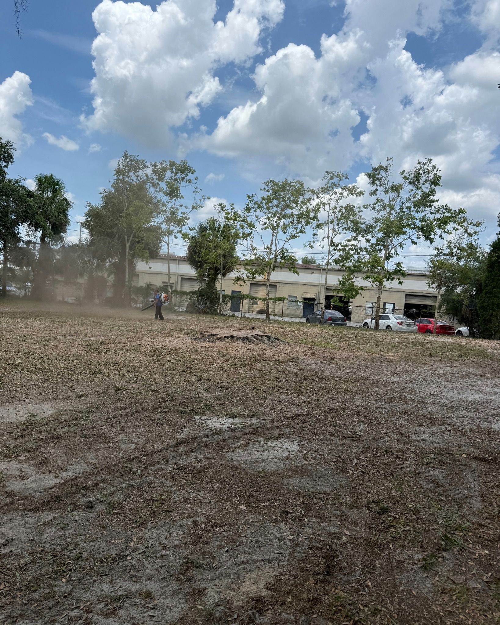 Dusty empty lot with trees, buildings, and parked cars under a cloudy blue sky
