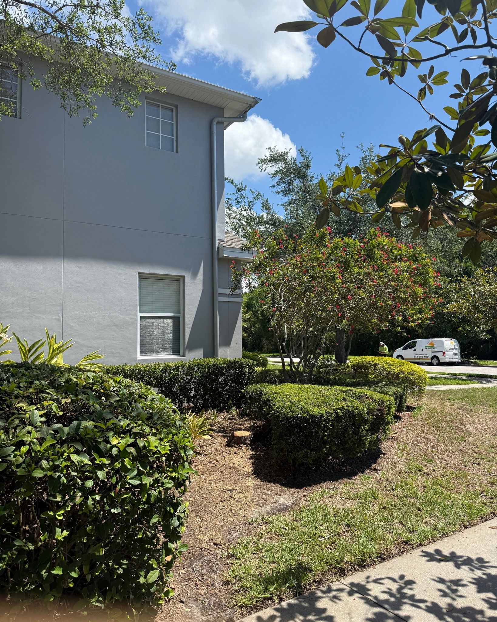 Apartment building and hedges beside a sidewalk, with trees and a parked white car in the background.