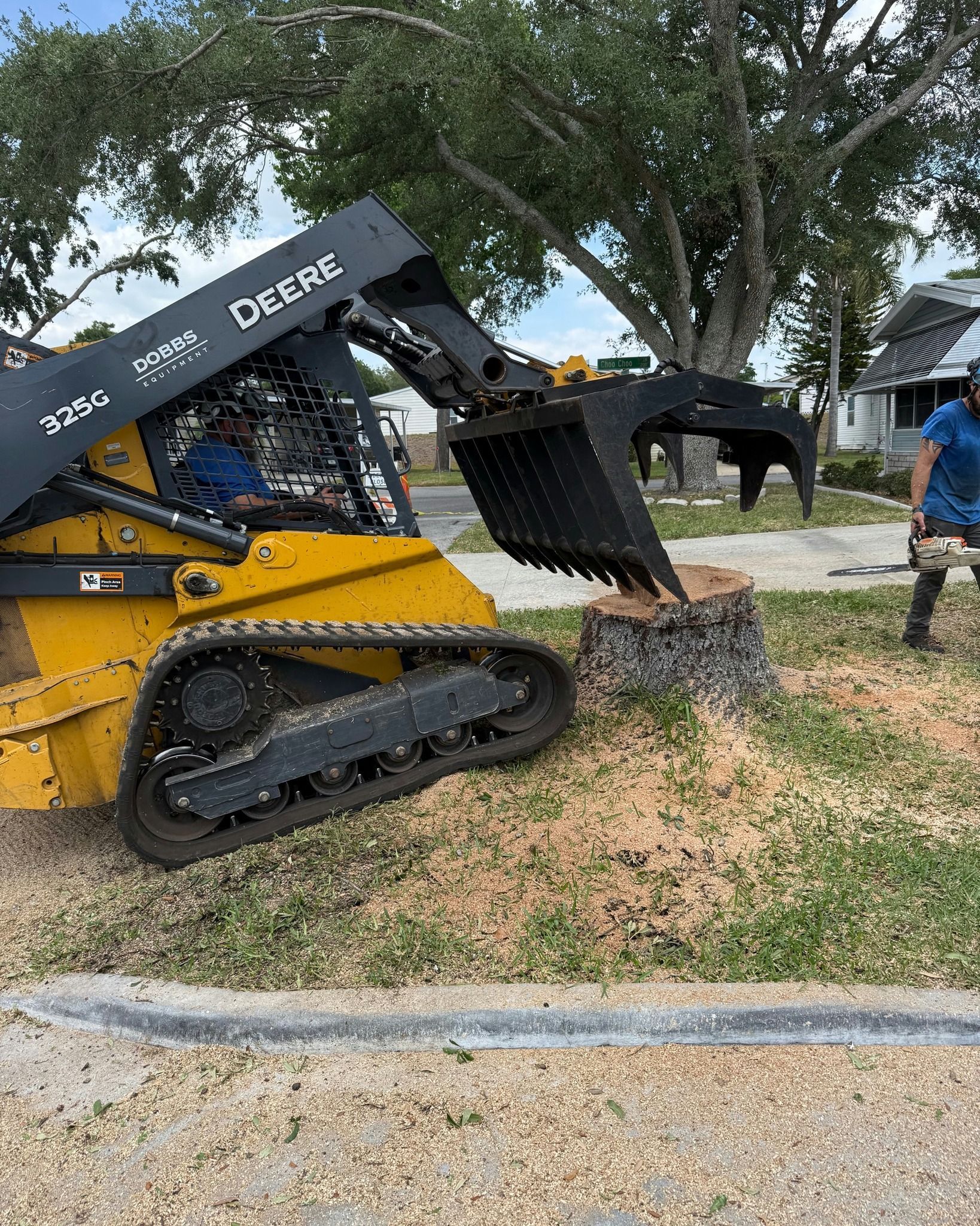 Yellow skid steer digging mulch beside a sidewalk, with trees and a person in blue nearby.