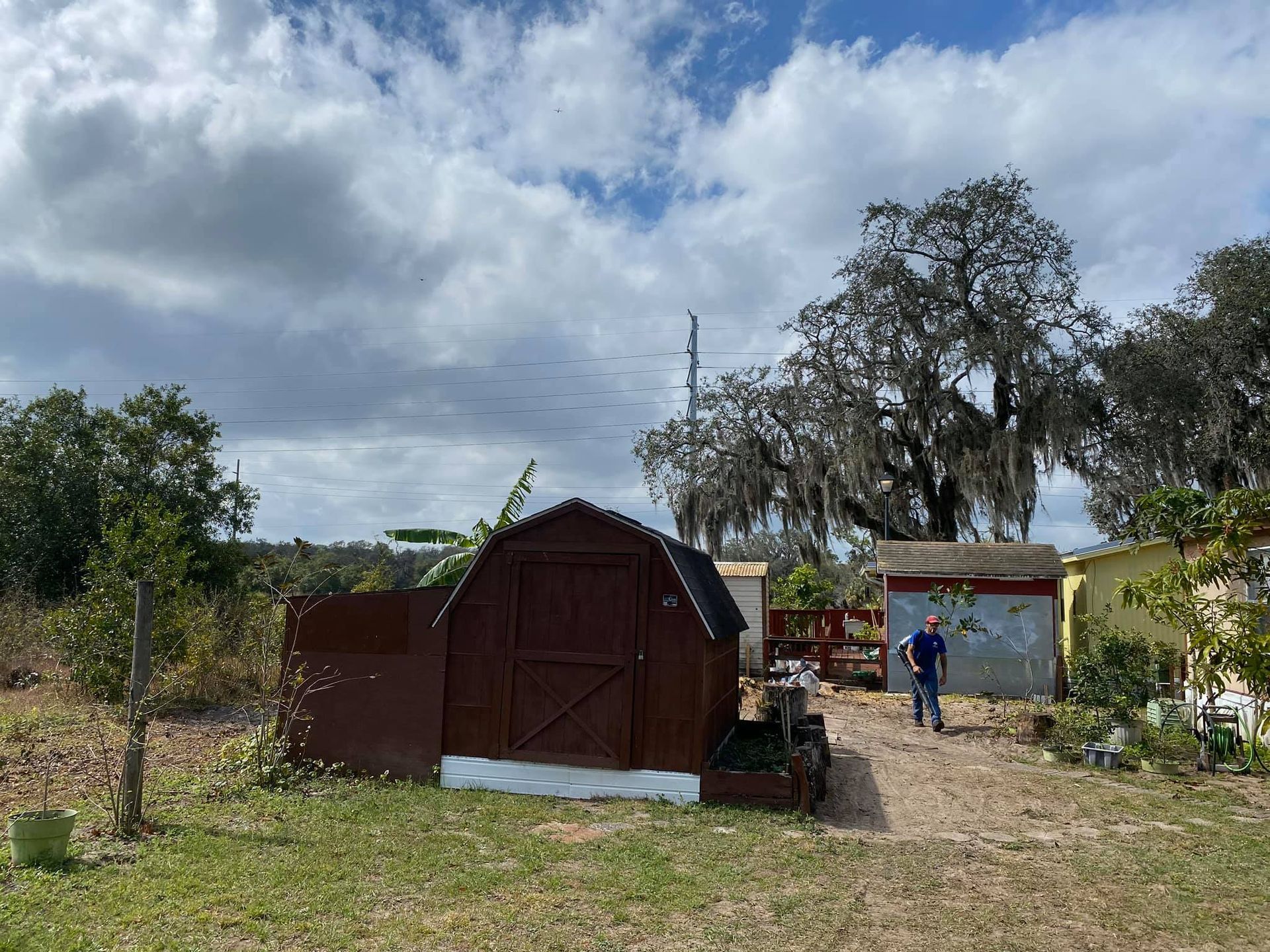 Rural yard with a brown shed, small building, and a person standing near a dirt path under cloudy sky