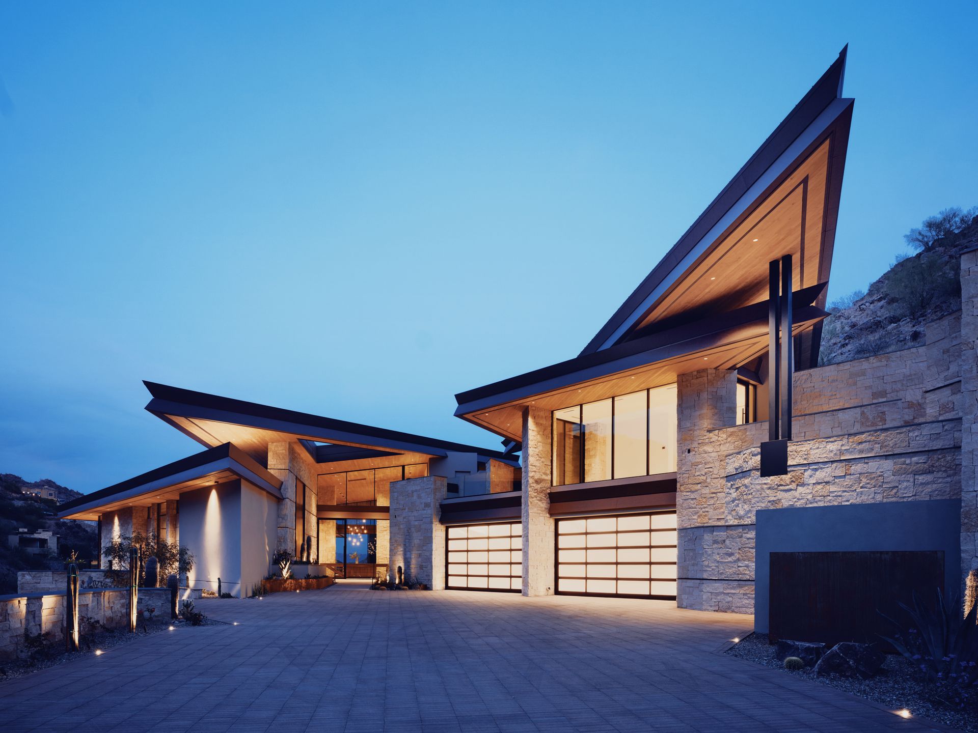 Modern home with angular roof, stone facade, glass garage doors, and a long driveway at dusk.