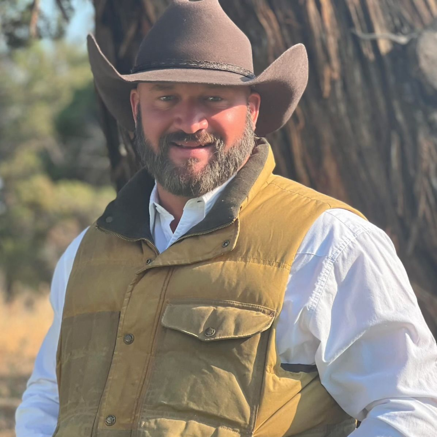 Smiling man in a brown cowboy hat and tan vest standing outdoors.