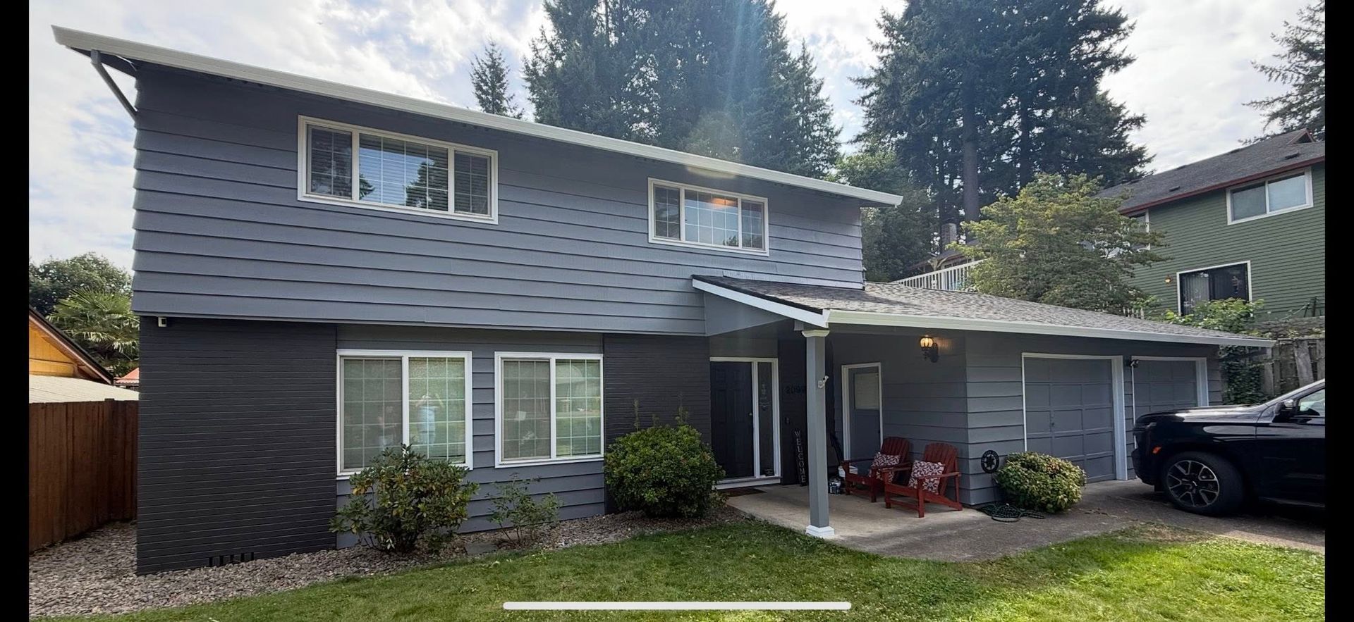 Two-story gray house with black lower siding, white-trimmed windows, and a covered porch in a wooded neighborhood