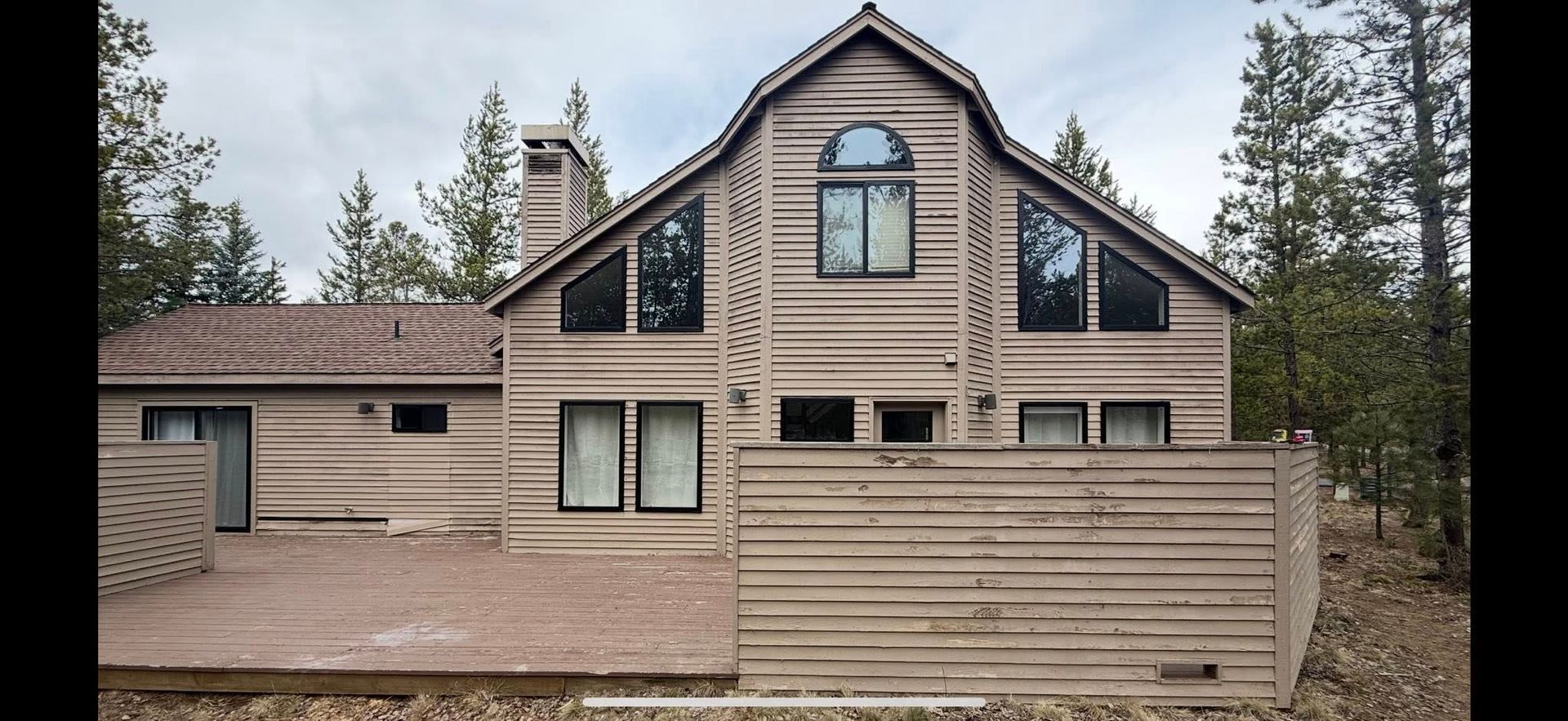 Log cabin-style house with beige siding and a wooden fence, surrounded by pine trees under a cloudy sky