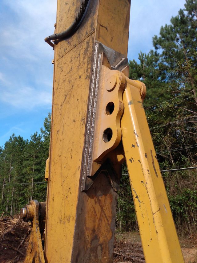 Close-up of a yellow excavator arm and hydraulic linkage against a blue sky and trees