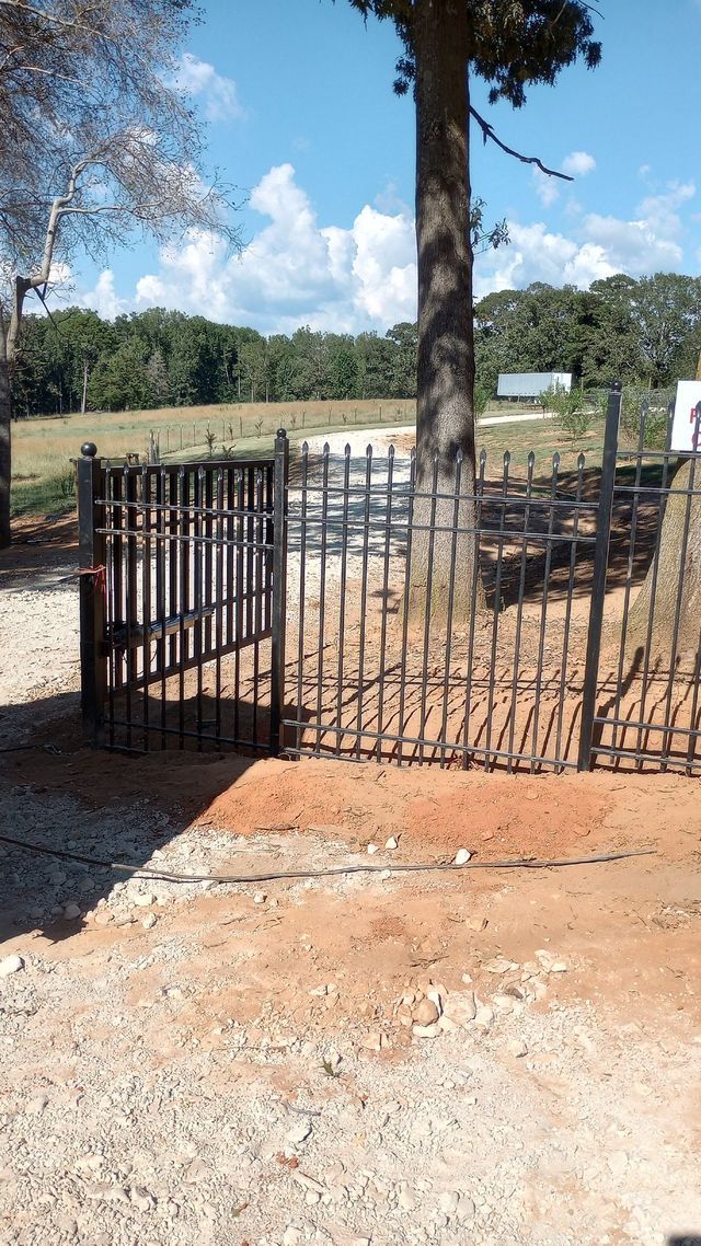 Black metal gate at a sunny rural entrance, with a dirt road, trees, and a field beyond.