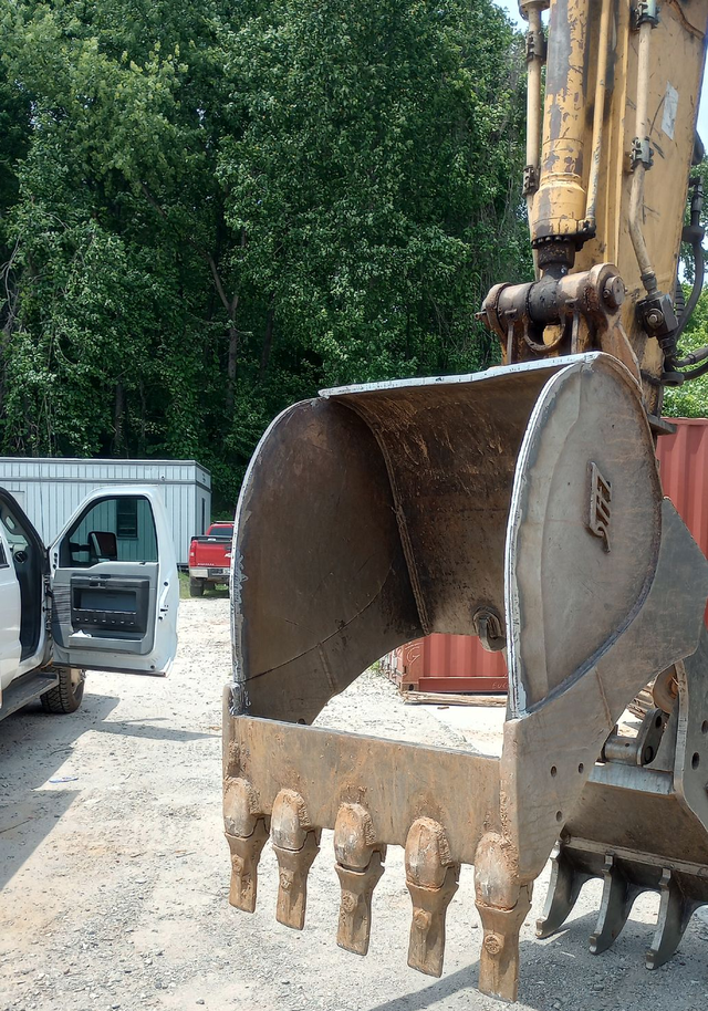 Excavator bucket with teeth at a construction site beside a white van and trees