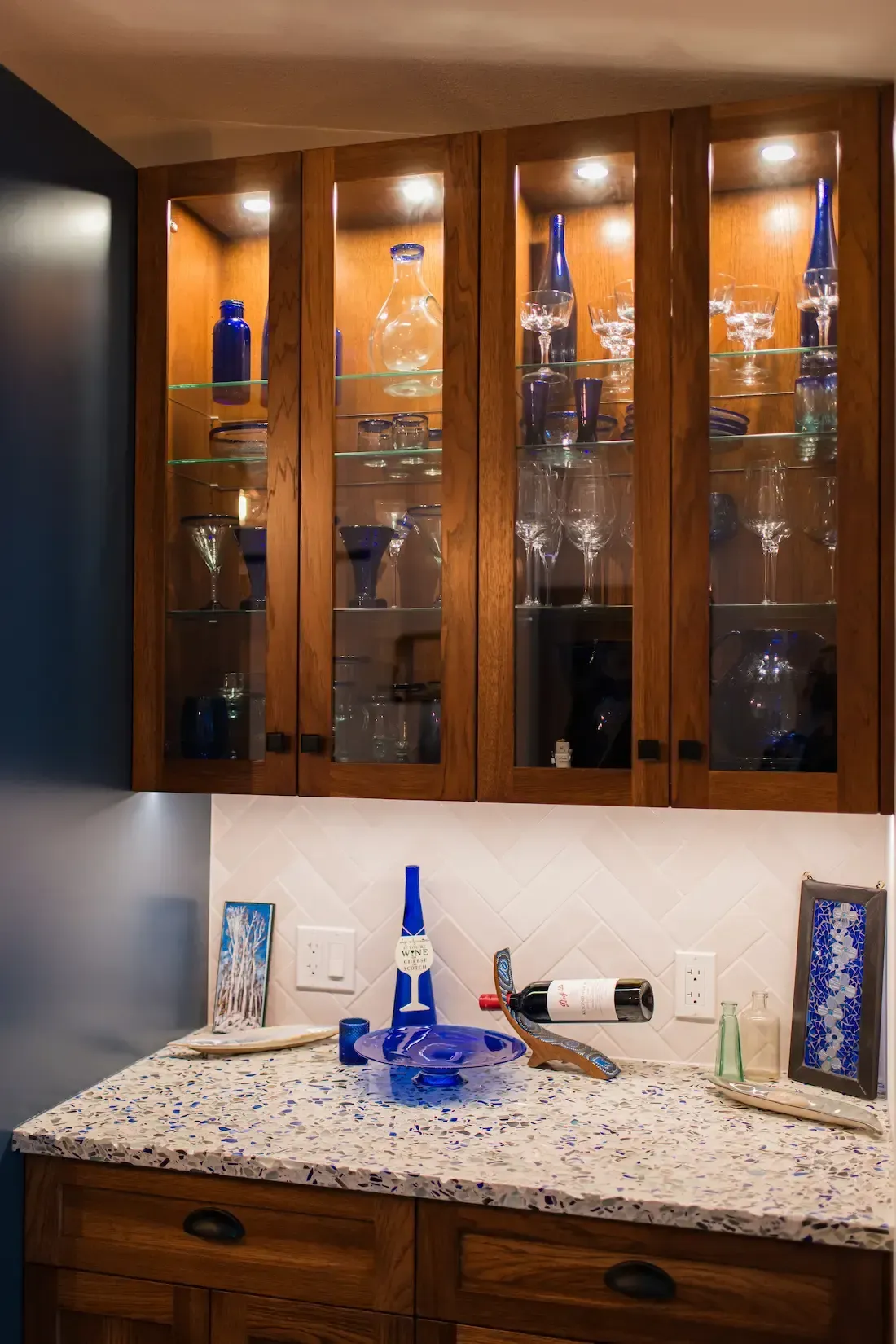 Wooden bar cabinet with glass shelves, blue glass decor, and a granite countertop sink under warm lighting