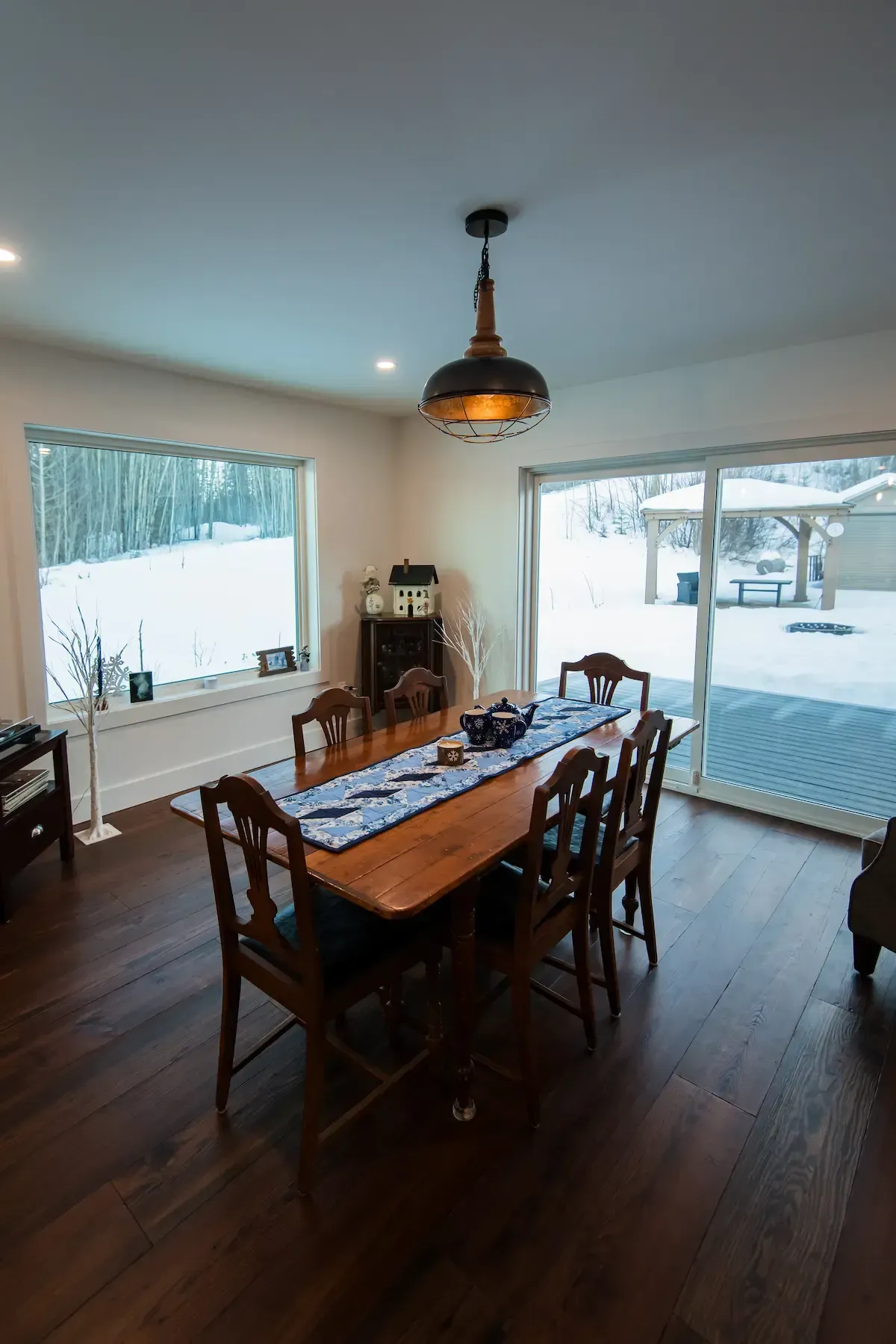 Dining room with wooden table and chairs, dark hardwood floor, and large windows overlooking a snowy yard.