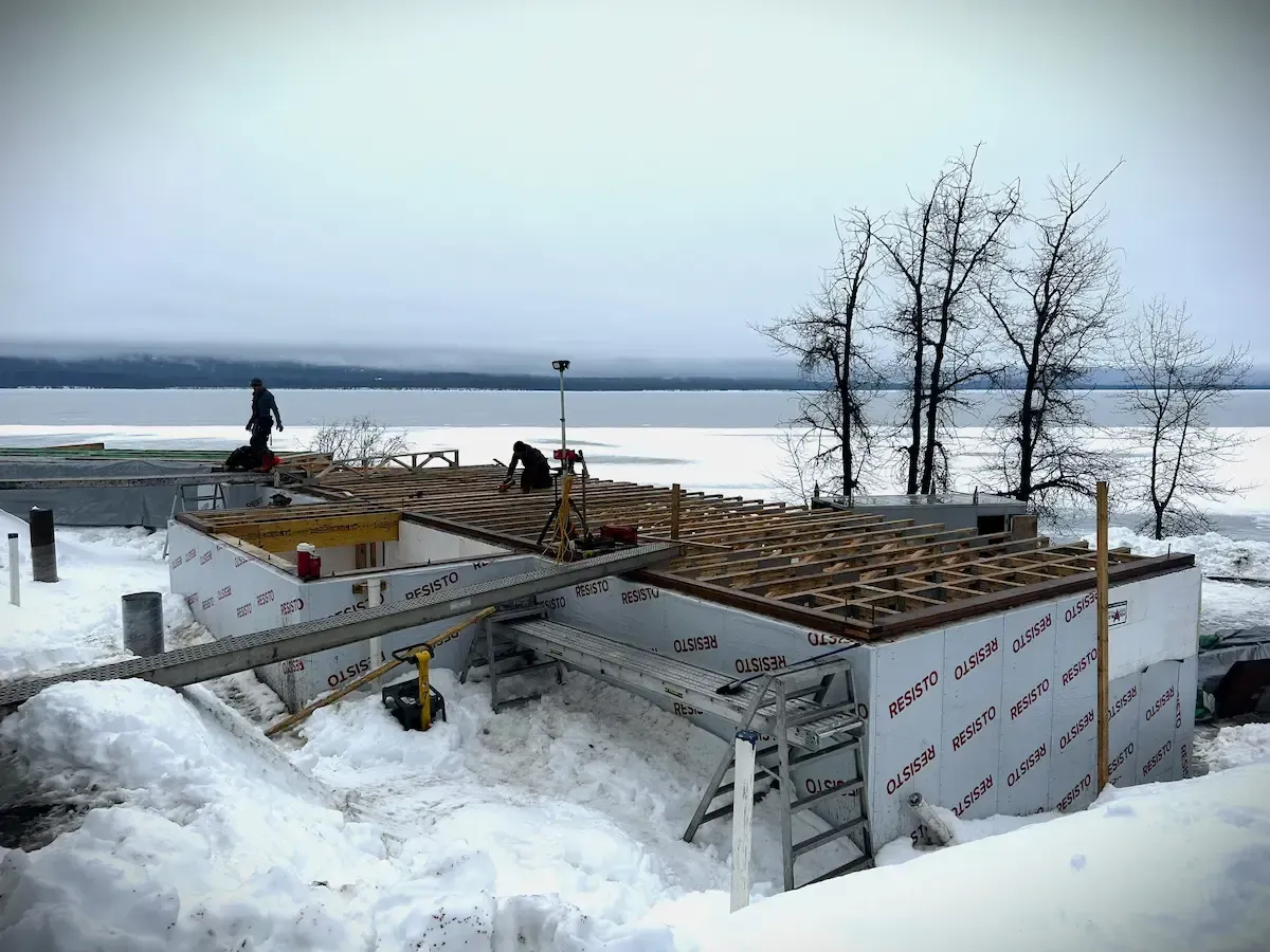 Snow-covered lakeside construction site with workers and a partially built wooden roof frame