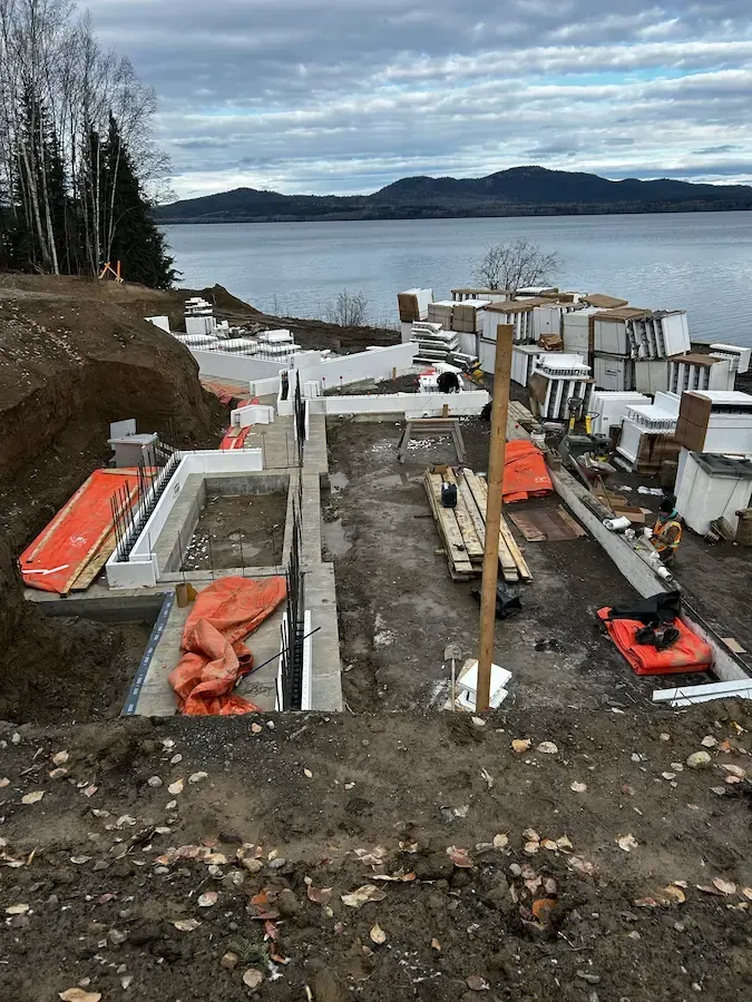 Construction site by a waterfront with concrete forms, equipment, and snow, overlooking a calm bay and mountains.
