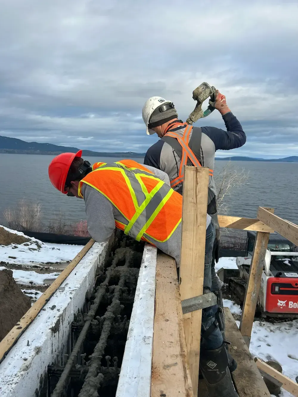 Two workers in safety gear repair a wooden dock beside calm water on a cloudy day.
