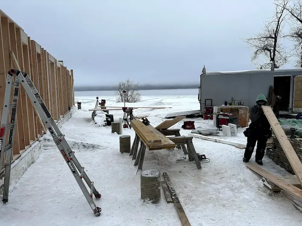 Snowy outdoor work area with ladders, benches, and a person near a shed by the water