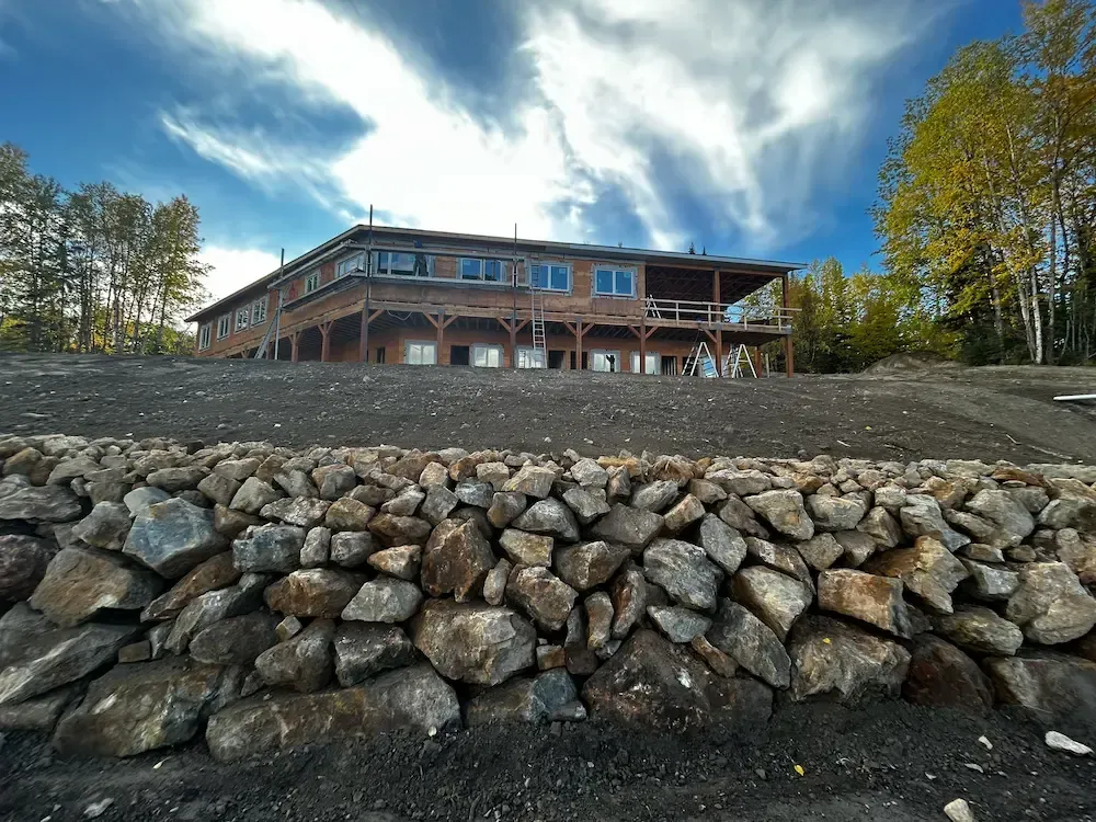 Large wooden building on a hill behind a stacked rock retaining wall under a cloudy blue sky