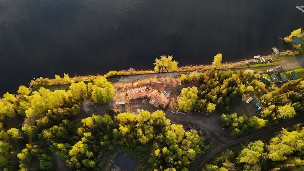 Aerial view of a rustic cabin beside a dark river, surrounded by dense green trees and golden foliage.