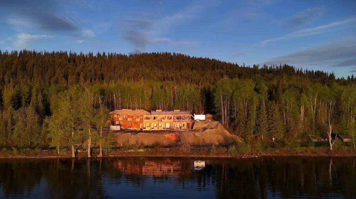 Lakeside cabins glowing in warm light at the edge of a forest, reflected in calm water at sunset