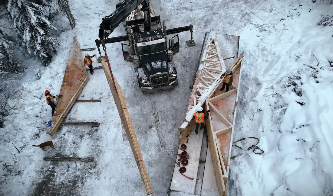 Snowy construction site with excavator lifting a long wooden beam beside a damaged bridge frame