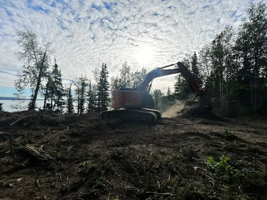 Excavator clearing a dirt lot in a forested area at sunset, with dust and tall trees in the background