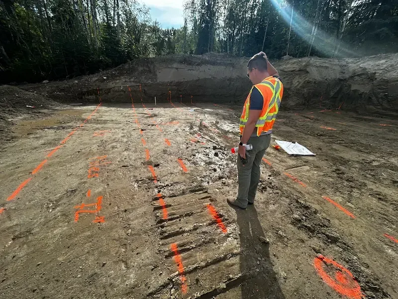 Worker in orange safety vest standing on marked dirt excavation site in wooded area