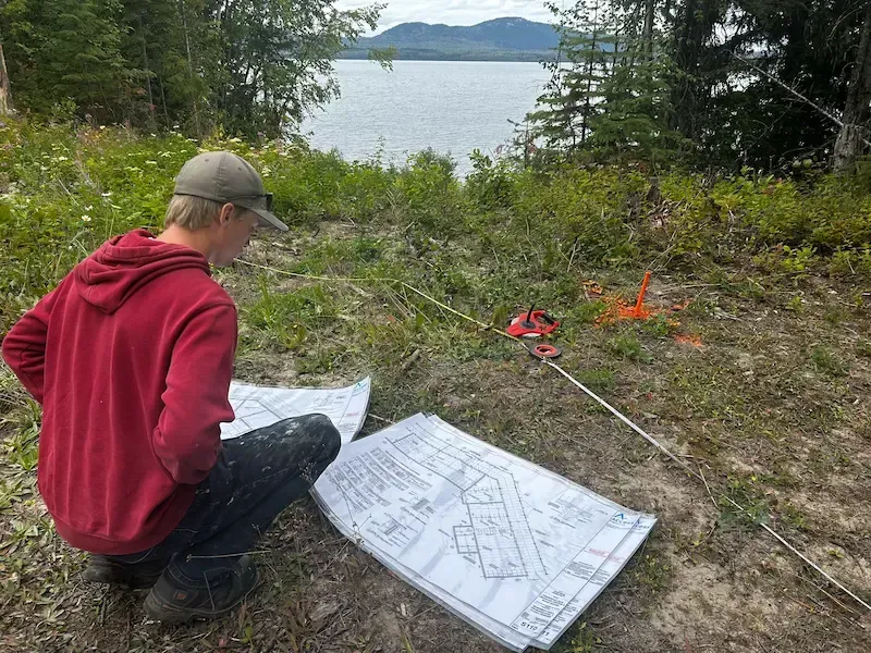 Person kneels over a map near a lake, setting survey markers in a wooded outdoor area.