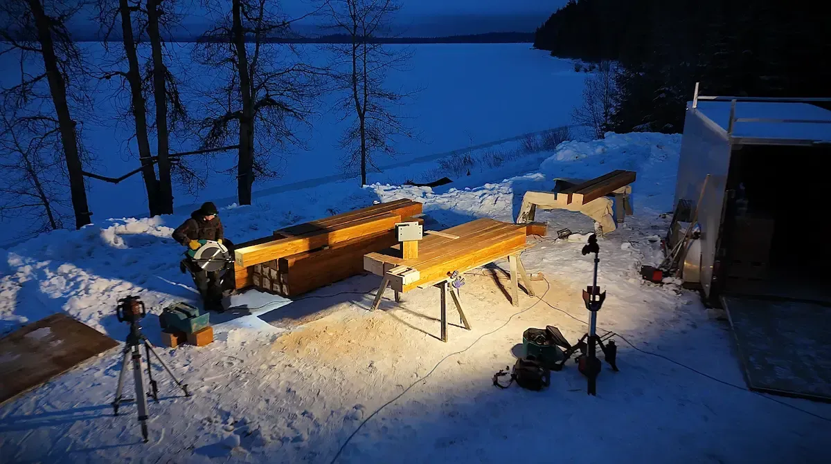 Snowy backyard at dusk with a lit wooden bench, lamp posts, and a small building by the water