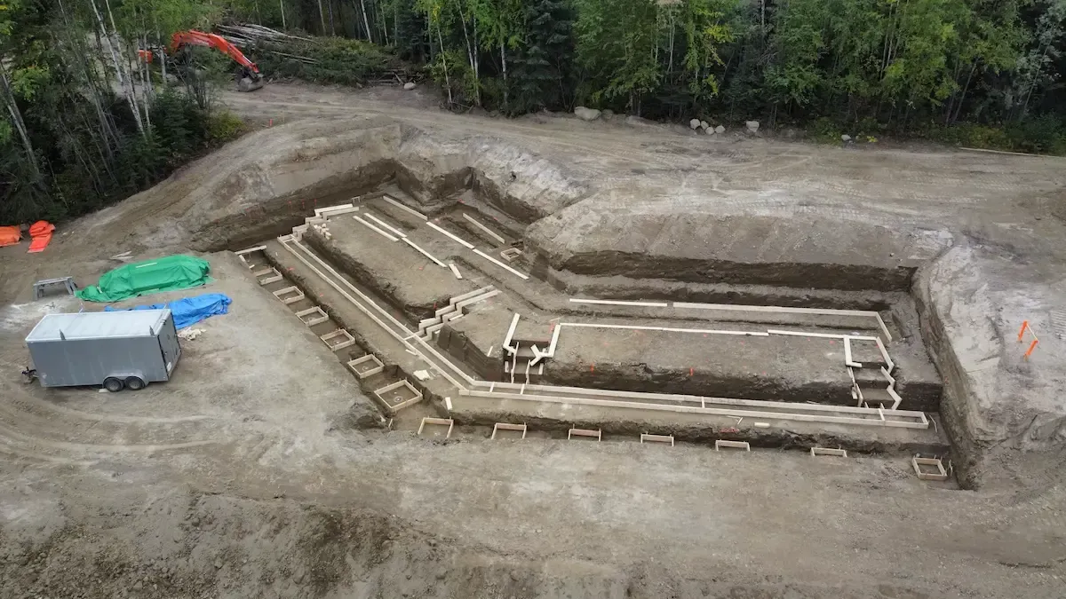 Excavated earthen foundation with concrete trench walls, viewed from above in a forest clearing