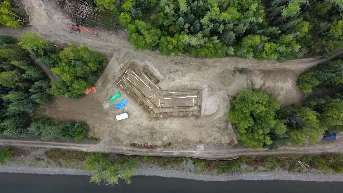 Aerial view of a dirt construction site surrounded by trees, with a small concrete structure and colorful materials.