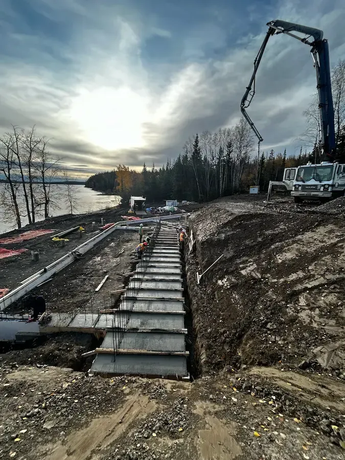 Construction site with a long walkway over dirt beside water, excavator arm raised at sunset.