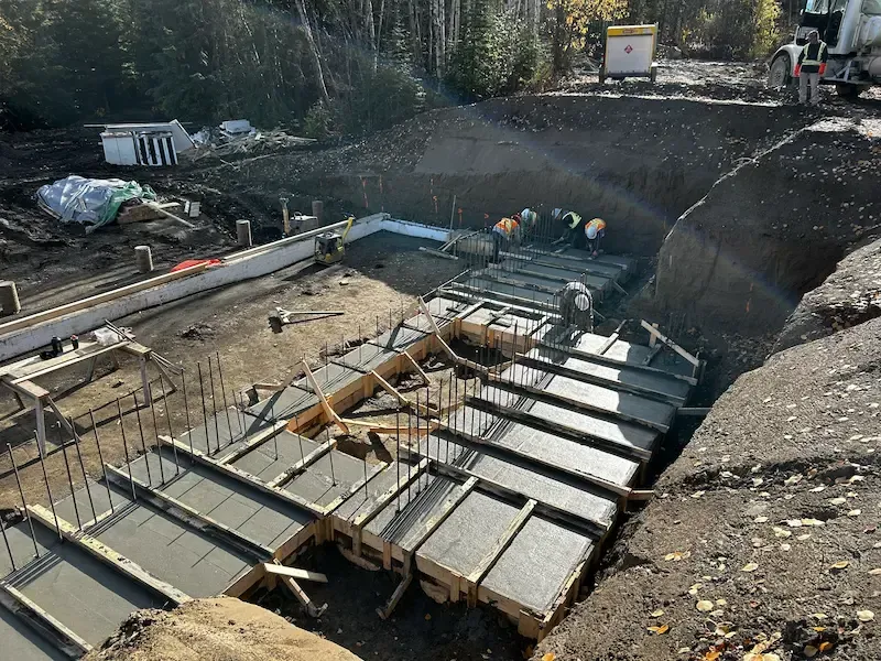 Construction site with concrete forms and rebar in a large excavation, workers in the background.
