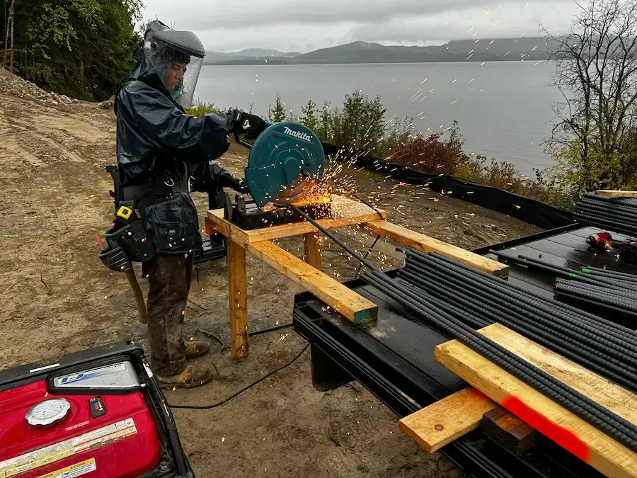 Person cutting metal pipes with a saw beside a rainy lakeside workbench