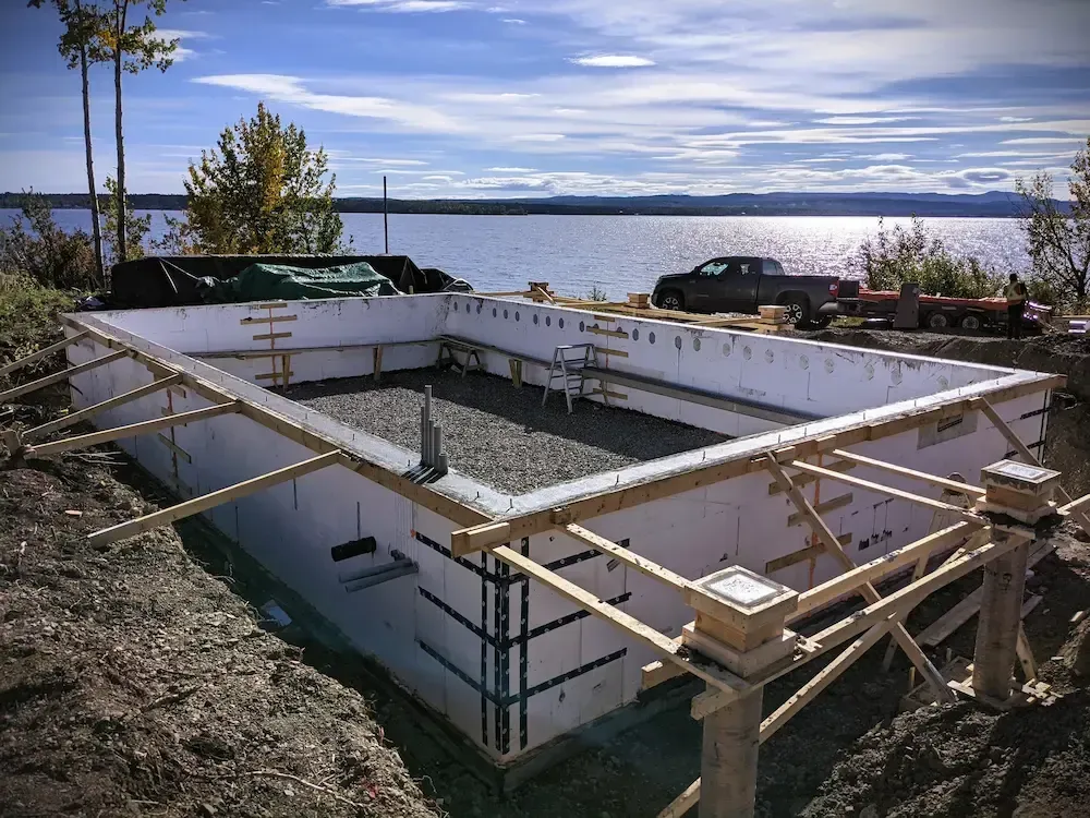 Construction site with a concrete foundation and wooden framing beside a lake under a cloudy sky
