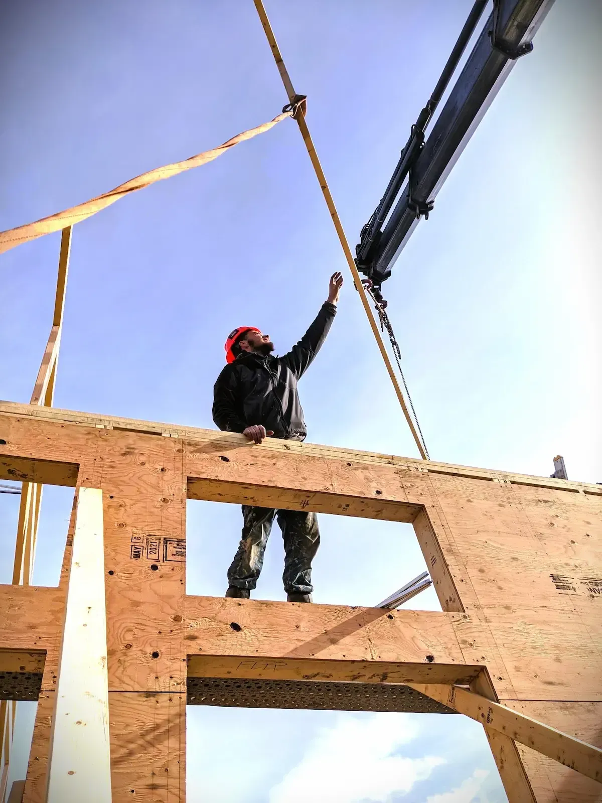 Construction worker in a hard hat lifting a beam on a wooden building frame against the sky